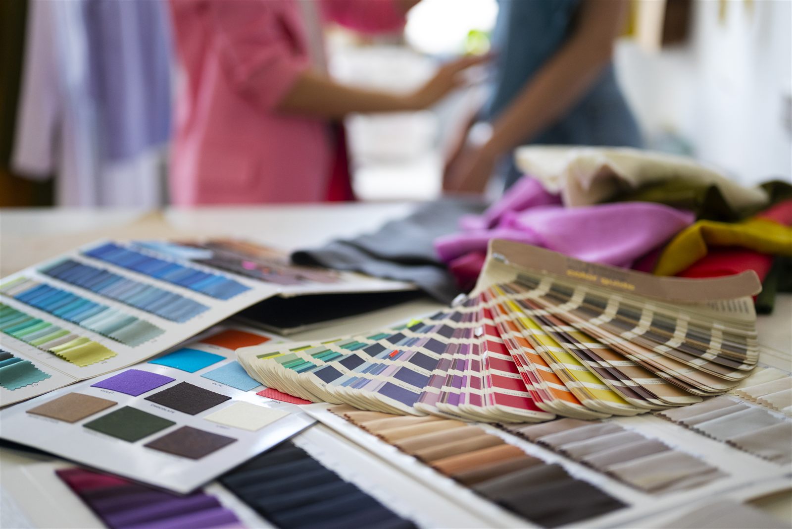 Muestrarios de colores y telas sobre una mesa de trabajo, con dos personas conversando al fondo